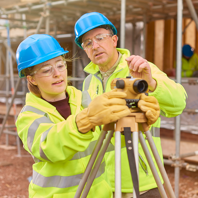 Two adults on a building site setting up a measuring tool on a tripod. 