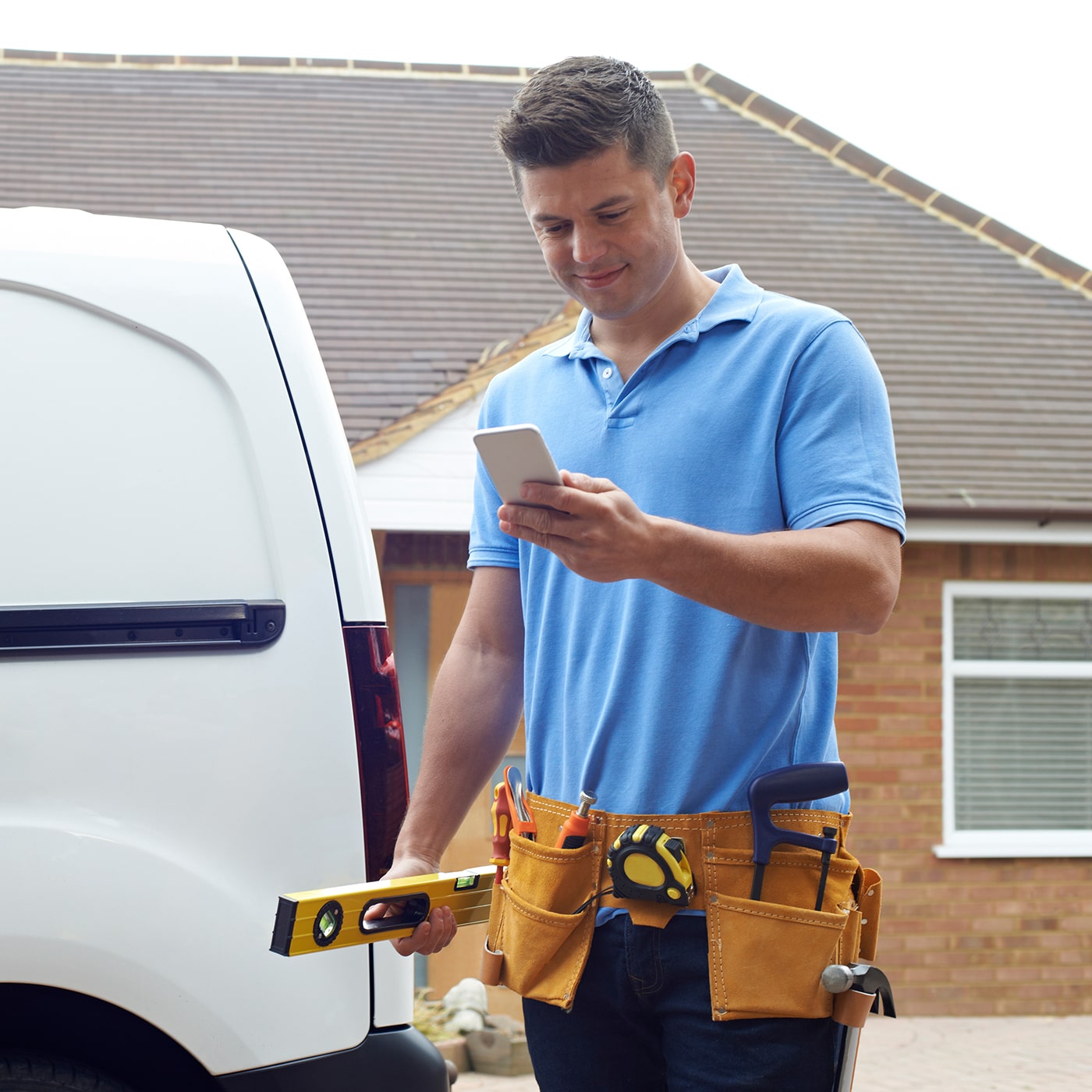 Builder wearing a toolbelt, standing beside his van and checking his mobile phone