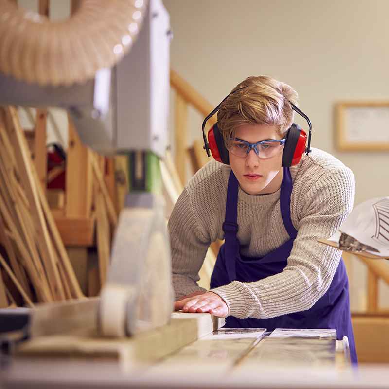 Tutor With Male Carpentry Student In Workshop Studying For Apprenticeship At College Using Bench Saw.